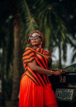 Elegant woman in vibrant striped outfit by a car with palm trees as background.